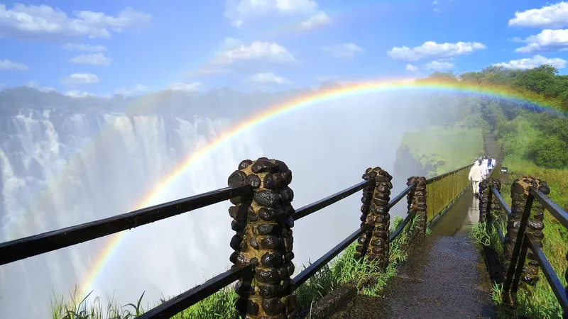 Rainbow over the falls from a wet footpath with stone-post railings, Zambian side