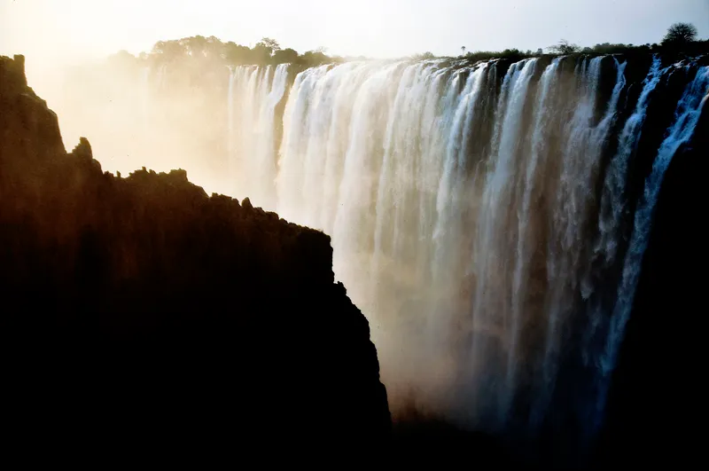 Falls against hazy morning light, a dark cliff in silhouette in front